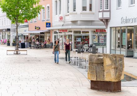 Zwei Frauen spazieren durch Lübbecke mit Geschäften und einem kleinen Brunnen.