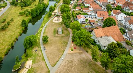 Luftaufnahme eines grünen Parks mit Spielplatz, umgeben von Fluss und roten Dächern der Stadt.
