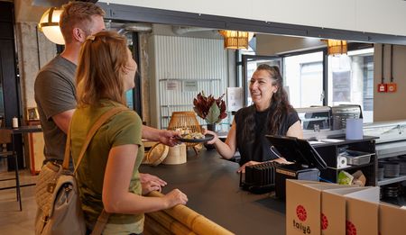 Drei Personen stehen an einer Theke in einem modernen Café, eine Serviererin lächelnd überreicht Snacks.