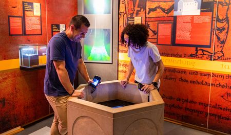 Zwei Personen betrachten im Museum einen historischen Taufstein, umgeben von Informationstafeln.