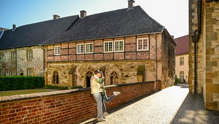 Ein Paar umarmt sich lachend auf einer Backsteinbrücke vor einer historischen Klosteranlage.