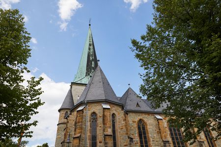 Ein historischer Kirchturm mit grünem Kupferdach, umgeben von üppigen Bäumen unter blauem Himmel.