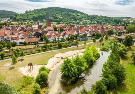 Luftaufnahme einer malerischen Stadt mit Fachwerkhäusern, Fluss und grüner Hügellandschaft im Hintergrund.