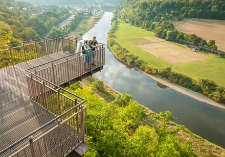 Menschen stehen auf einer Aussichtsplattform mit Blick auf einen Fluss und bewaldete Hügel.