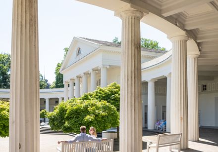 Klassizistische Architektur mit weißen Säulen in einem sonnigen Innenhof; Menschen auf einer Bank.