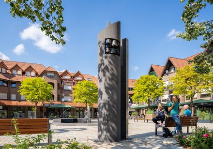 Ein moderner Glockenturm auf einem belebten Platz mit Sitzbänken und entspannten Menschen an einem sonnigen Tag.
