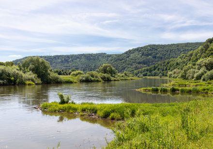 <alt>Ruhige Flusslandschaft mit grünen Ufern und bewaldeten Hügeln unter blauem Himmel.</alt>