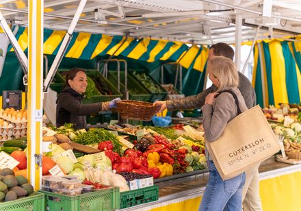 Marktszene mit paarigem Kunden vor einem farbenfrohen Verkaufsstand, reich an frischem Obst und Gemüse.
