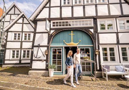 Fachwerkhaus mit grünem Tor und goldenem Anker, Paar geht Hand in Hand auf Kopfsteinpflaster vorbei