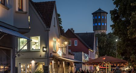 Abendliche Straßenszene mit beleuchtetem Wasserturm, Cafés, Restaurants und Flanieren in gemütlicher Atmosphäre.