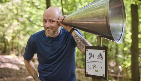 Mann lauscht im Leistruper Wald einem Waldhörrohr. Umgeben von grünen Bäumen und Tageslicht.