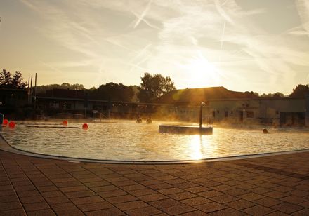 Ein beleuchtetes Freibad mit Sprüngen im Wasser und dunklen Silhouetten bei Sonnenuntergang.