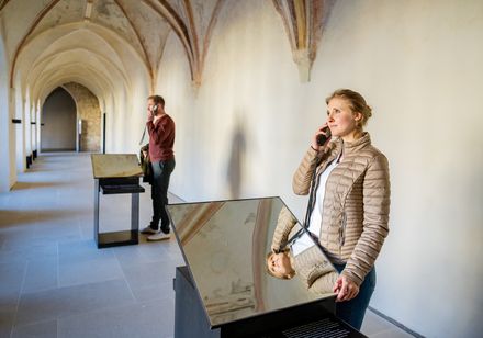 Zwei Besucher erkunden eine Ausstellung im gotischen Gang eines historischen Gebäudes mit Audioguides.
