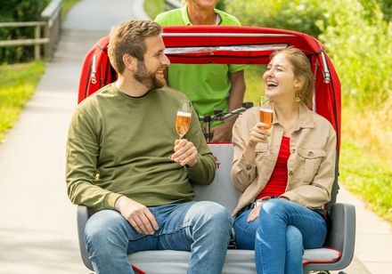 Ein Paar genießt lächelnd eine Fahrt in einer Rikscha mit einem älteren Fahrer auf einem sonnigen Weg.
