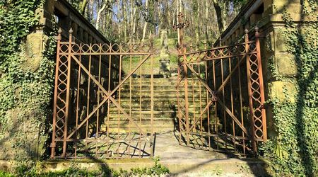 Historisches Metalltor führt zu einer von Bäumen umrahmten steinernen Treppe in einem bewaldeten Park.