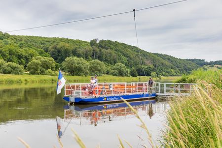 <p>Eine kleine Seilfähre überquert einen ruhigen Fluss, umgeben von grüner Landschaft und Bäumen.</p>