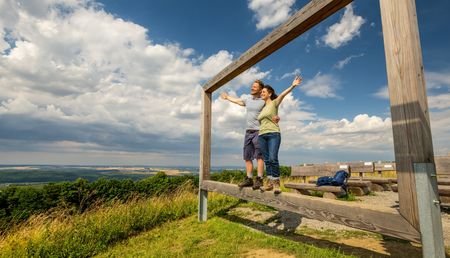 Zwei fröhliche Wanderer posieren in einem hölzernen Bilderrahmen vor einer weiten Landschaft.