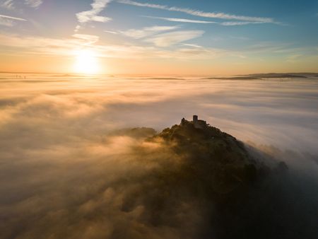 Ruine auf nebelverhülltem Hügel bei Sonnenaufgang, umgeben von sanften Wolken und klarer Horizont.