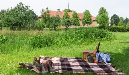 Picknickdecke mit Korb und Schuhen auf ländlicher Wiese, im Hintergrund Bäume und ein Gebäude.