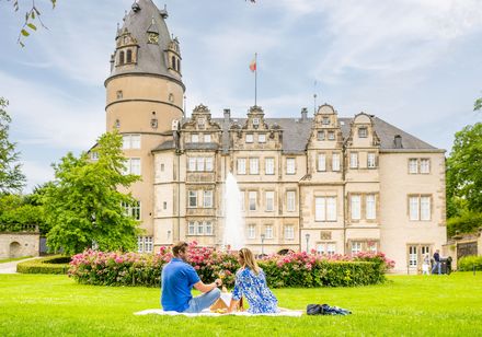 Zwei Personen entspannen auf einer Wiese vor dem historischen Schloss Detmold mit Turm und Brunnen.