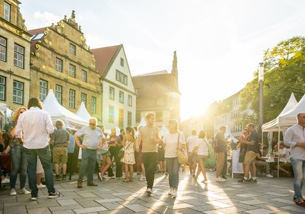 Menschen genießen Sonnenuntergang auf einem belebten Platz mit historischen Gebäuden und weißen Zelten.