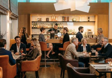 Modern gestalteter Hotelbar-Bereich mit sitzenden Gästen an Tischen und Barhockern, während ein Barkeeper Getränke vor einem beleuchteten Regal zubereitet.