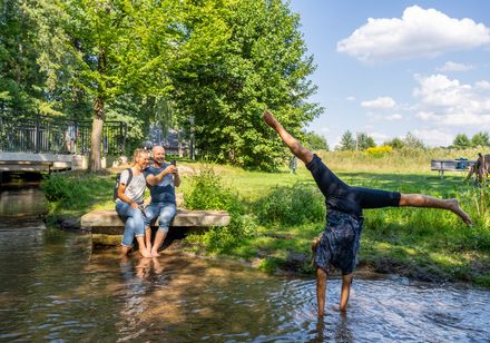 Familie sitzt fröhlich am Ufer eines Bachs, während ein Kind spielerisch einen Handstand macht.