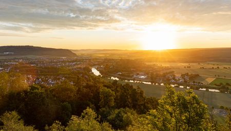 Sonnenuntergang über einer idyllischen Flusslandschaft im Tal mit Feldern und Hügeln im Hintergrund.