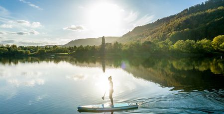 Person beim Stand-Up-Paddling auf einem ruhigen See im Sonnenuntergang, umgeben von bewaldeten Hügeln.
