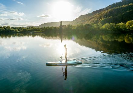 Person beim Stand-Up-Paddling auf einem ruhigen See im Sonnenuntergang, umgeben von bewaldeten Hügeln.