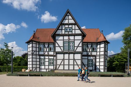 Fachwerkhaus mit rotem Ziegeldach und weißer Fassade, drei Personen gehen davor entlang.