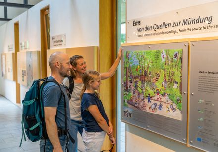 Familie betrachtet eine Wandtafel mit buntem Illustrationsbild in einem Besucherzentrum.