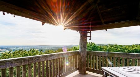 Holzplattform mit Geländer, Sonnenstrahlen brechen durch das Dach, Panoramablick auf weitläufige Landschaft.