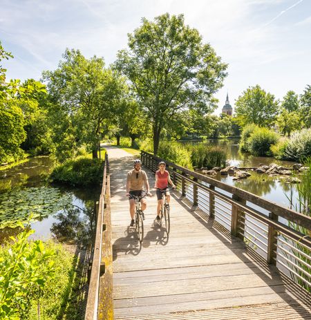 Zwei Radfahrer auf einem Holzsteg in Rheda-Wiedenbrück, im Hintergrund ein schlanker Kirchturm.