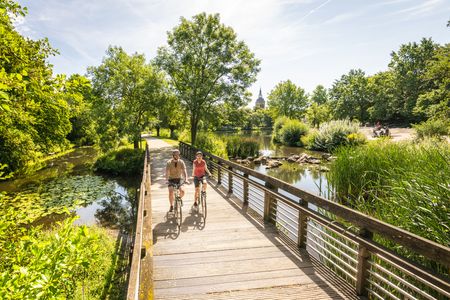 Zwei Radfahrer auf einem Holzsteg in Rheda-Wiedenbrück, im Hintergrund ein schlanker Kirchturm.