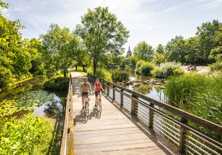 Zwei Radfahrer auf einem Holzsteg in Rheda-Wiedenbrück, im Hintergrund ein schlanker Kirchturm.