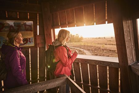 Zwei Frauen im Holzbeobachtungsturm betrachten den Sonnenuntergang über einer weitläufigen Landschaft.