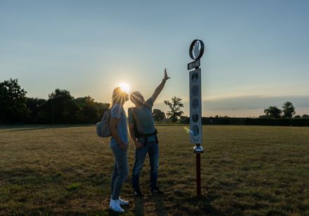 Zwei Personen stehen auf einer Wiese und betrachten gemeinsam eine Infotafel im Abendlicht.