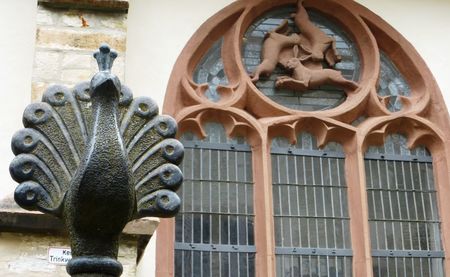 Steinernes Pfauenrelief vor kunstvollem gotischen Fenster mit Figuren von kämpfenden Hasen.