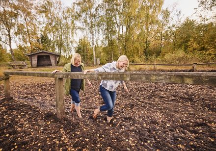 Zwei Frauen gehen lachend barfuß durch einen matschigen Parcours, umgeben von Herbstbäumen.