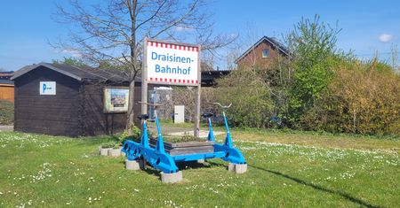 Eine blaue Draisine steht auf einem Rasenstück vor einem Schild mit der Aufschrift „Draisinen-Bahnhof“.