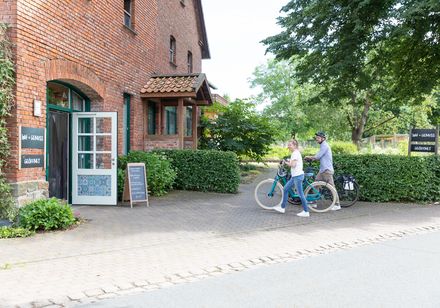 Rotes Backsteinhaus, geöffnete Tür, zwei Radfahrer im Vordergrund auf einem gepflasterten Weg.