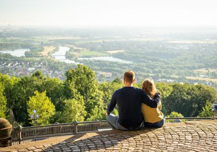 Ein Paar sitzt auf einer Mauer und blickt in eine weitläufige, grüne Landschaft mit Fluss.
