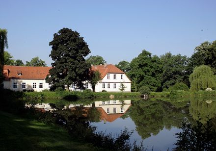 Ein Herrenhaus am Teichufer, umgeben von Bäumen, spiegelt sich im ruhigen Wasser wider.