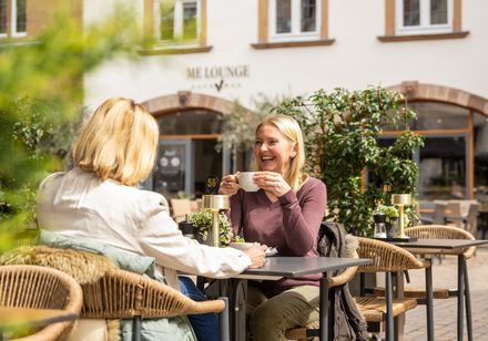 Zwei Frauen sitzen fröhlich bei Kaffee im Freien an einem Café-Tisch in einer belebten Fußgängerzone.