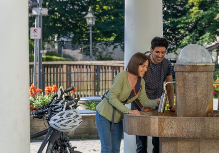 Zwei Personen füllen Trinkflaschen an einem öffentlichen Brunnen auf, Fahrräder stehen daneben.