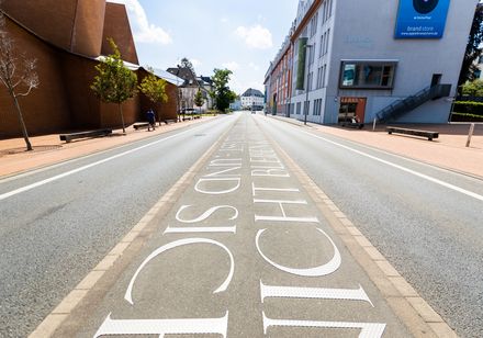 Breite Straße mit Kunstinstallation: "ICH" und "FÜR SICH" in großen Buchstaben auf Asphalt.