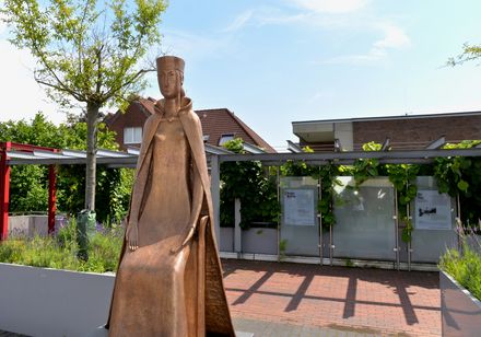 Bronzestatue einer sitzenden Frau in mittelalterlicher Kleidung, umgeben von grüner Bepflanzung.