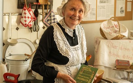 Ältere Frau im traditionellen Kleid, lächelnd in einer nostalgischen Küche mit historischen Requisiten.