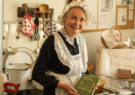 Ältere Frau im traditionellen Kleid, lächelnd in einer nostalgischen Küche mit historischen Requisiten.